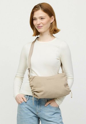 Junge Frau mit kurzen roten Haaren, die ein weißes Oberteil und Jeans trägt und eine beige Stoff-Umhängetasche quer über den Körper trägt.