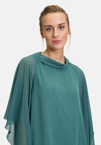 Top in chiffon di colore teal con scollo a cascata e maniche fluenti. Presenta un tessuto leggero e trasparente che si adagia delicatamente attorno al corpo.