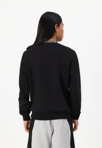 Juoda crewneck tipo megztinis su įaudžiamais rankogaliais ir apatine kraštine, ant nugaros apykaklės šviečia balta "LACOSTE" logotipas. Minkšta medžiaga, laisva prigludimo forma.