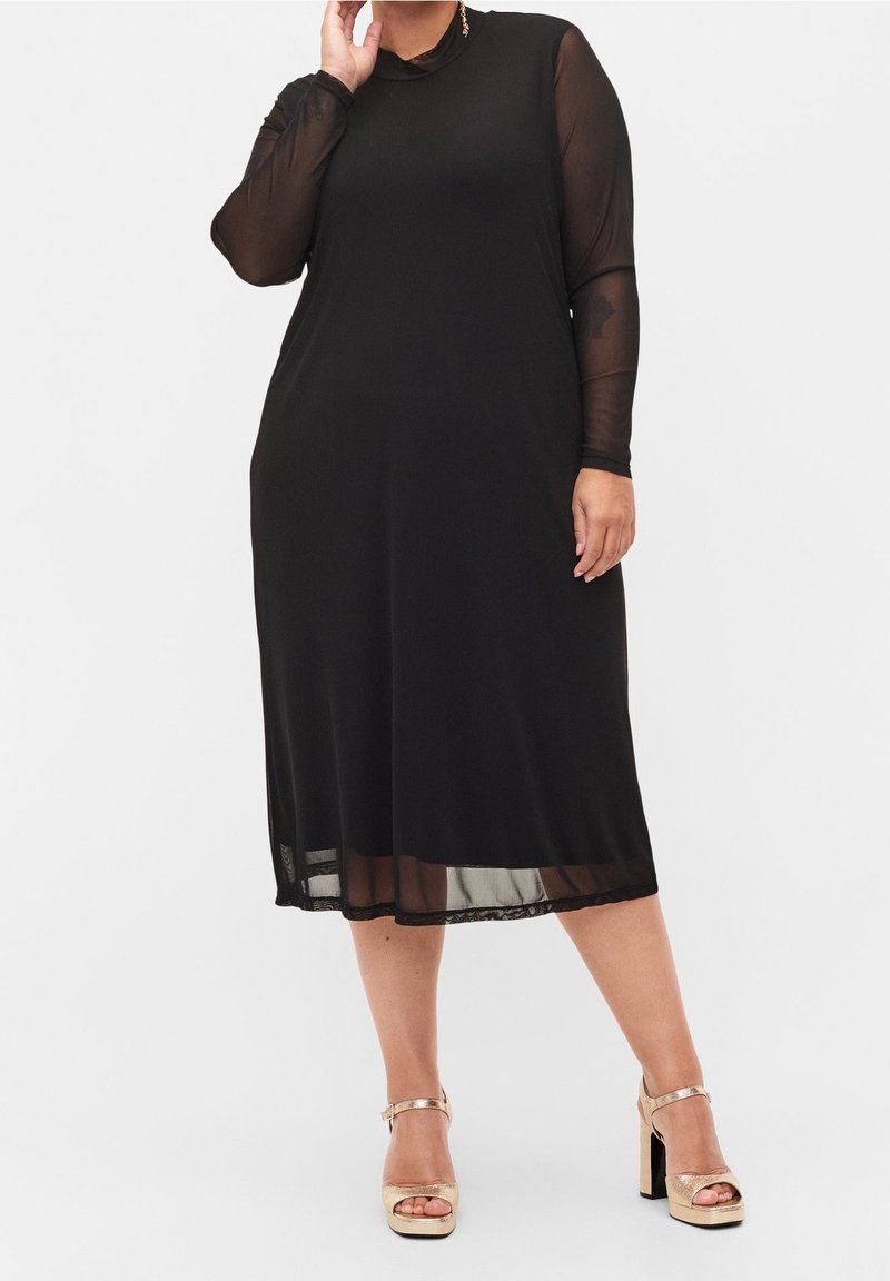 Femme portant une robe midi noire avec des manches longues en tulle transparent et des sandales à talons hauts dorées, debout devant un fond blanc.