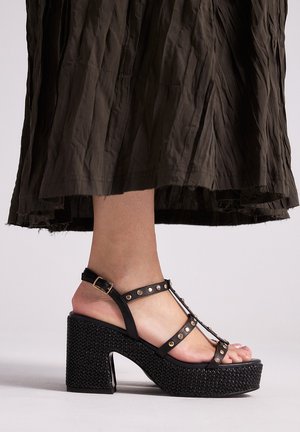 Pie femenino con sandalia de tacón bloque negro con plataforma, adornada con tachuelas doradas y correa en el tobillo, combinada con una falda arrugada de color marrón oscuro.