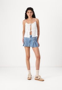 Witte mouwloze top met strik aan de voorkant en oogjesdetails, gecombineerd met een blauwe denim plissé mini-rok en beige sneakers met een patroon.