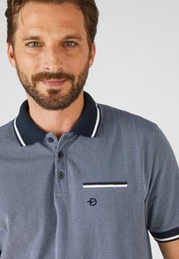 Polo da uomo di colore blu scuro con un motivo testurizzato, colletto a righe, pattina con tre bottoni e una tasca decorativa.