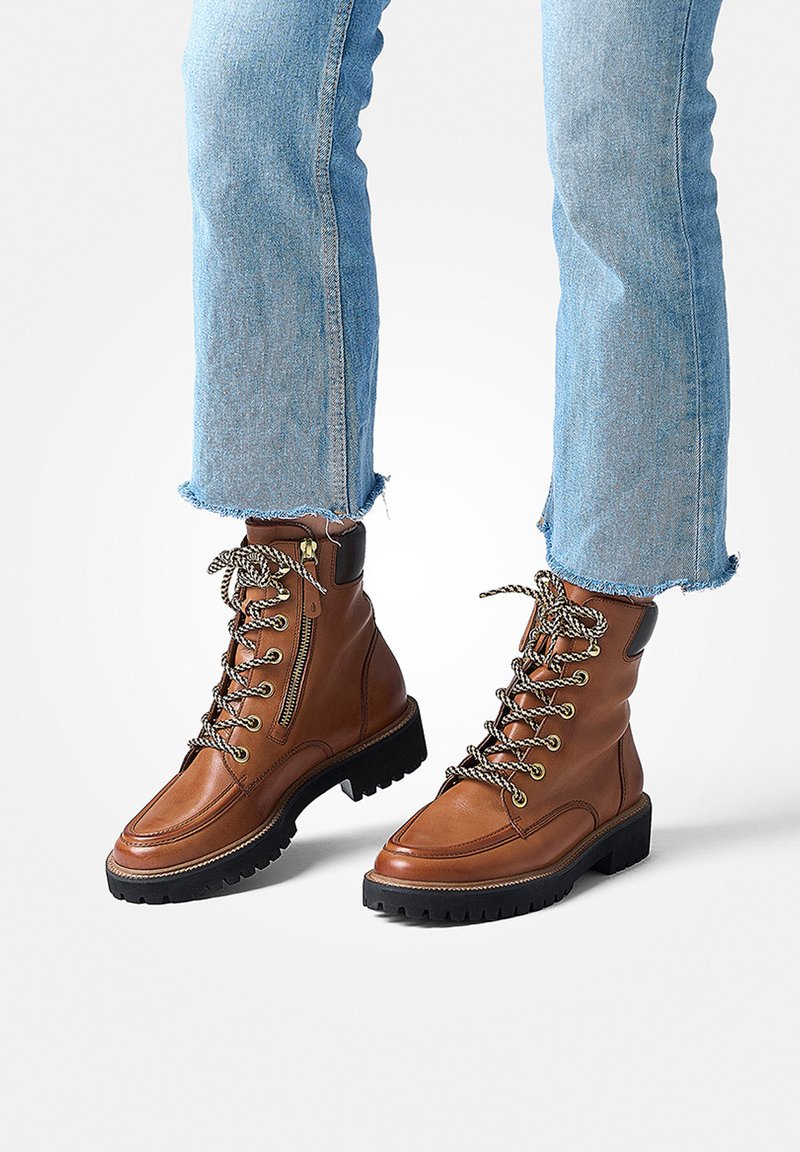 Bottes chelsea en cuir marron avec des lacets texturés, des accents en métal et une semelle en caoutchouc noir robuste. Assorties à un jean en denim léger bleu avec des bords effilochés.