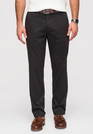 Mężczyzna noszący czarne proste spodnie, brązowy pasek, brązowe skórzane mokasyny, białą prążkowaną koszulę z krótkim rękawem i srebrny zegarek na rękę.