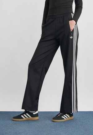 Personne portant un pantalon de survêtement Adidas noir avec des bandes blanches et des baskets Adidas noires, debout les mains dans les poches devant un fond uni.