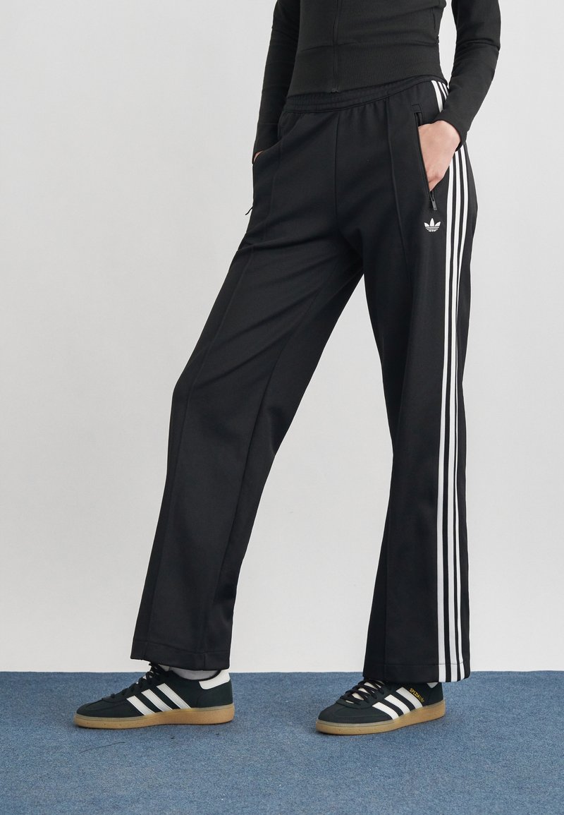 Personne portant un pantalon de survêtement Adidas noir avec des bandes blanches et des baskets Adidas noires, debout les mains dans les poches devant un fond uni.