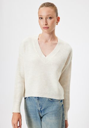 Cremefarbener Rippstrickpullover mit V-Ausschnitt und langen Ärmeln, in lässiger Passform und aus weichem Material, kombiniert mit hellblauen Jeans.