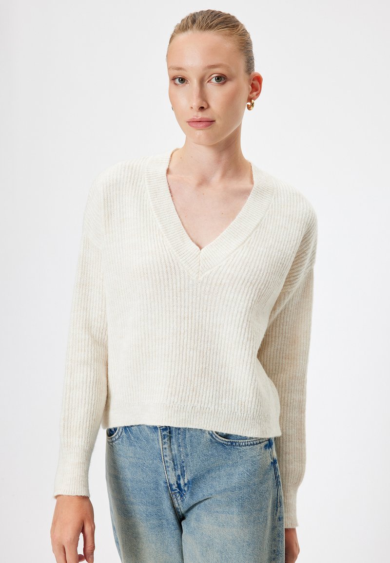 Maglione a coste color avorio con scollatura a V e maniche lunghe, caratterizzato da una vestibilità comoda e una texture morbida, abbinato a jeans azzurri chiari.