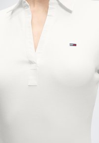 Λευκό ριμπ πλεκτό πόλο με ανοιχτό γιακά και μικρό κεντημένο λογότυπο σε σκούρο μπλε, κόκκινο και λευκό στο αριστερό στήθος.