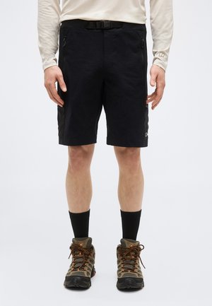 Henkilö, jolla on mustat ulkoilushortsit, beige pitkähihainen paita, mustat sukat ja ruskeat vaelluskengät, seisomassa yksinkertaista taustaa vasten.