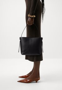 Borsa a secchiello in pelle nera con un manico corto, texture liscia e design minimalista. Il modello indossa un abito marrone a coste e tacchi a punta marroni.