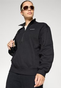 Maglione in fleece nero con colletto a mezzo zip, polsini e orlo a coste; presenta un logo sul lato sinistro del petto. Indossato con occhiali da sole.