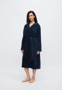 Μπλε σκούρο μπουρνούζι από πετσέτα με κουκούλα, μακριά μανίκια, ζώνη και δύο μπροστινές τσέπες. Μαλακή υφή, μονόχρωμο και με ελάχιστο λογότυπο.