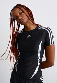 Young woman with long red braids wearing a black Adidas sports shirt with white stripes and standing against a plain light background.