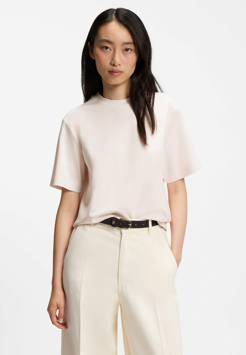 Femme aux longs cheveux noirs portant une chemise à manches courtes beige clair rentrée dans un pantalon crème taille haute avec une ceinture noire.