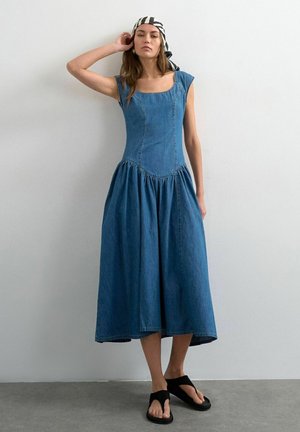 Kvinde iført en ærmeløs blå denim midikjole, sorte sandaler og et sort-hvidt mønstret tørklæde, der står mod en ensfarvet hvid væg.