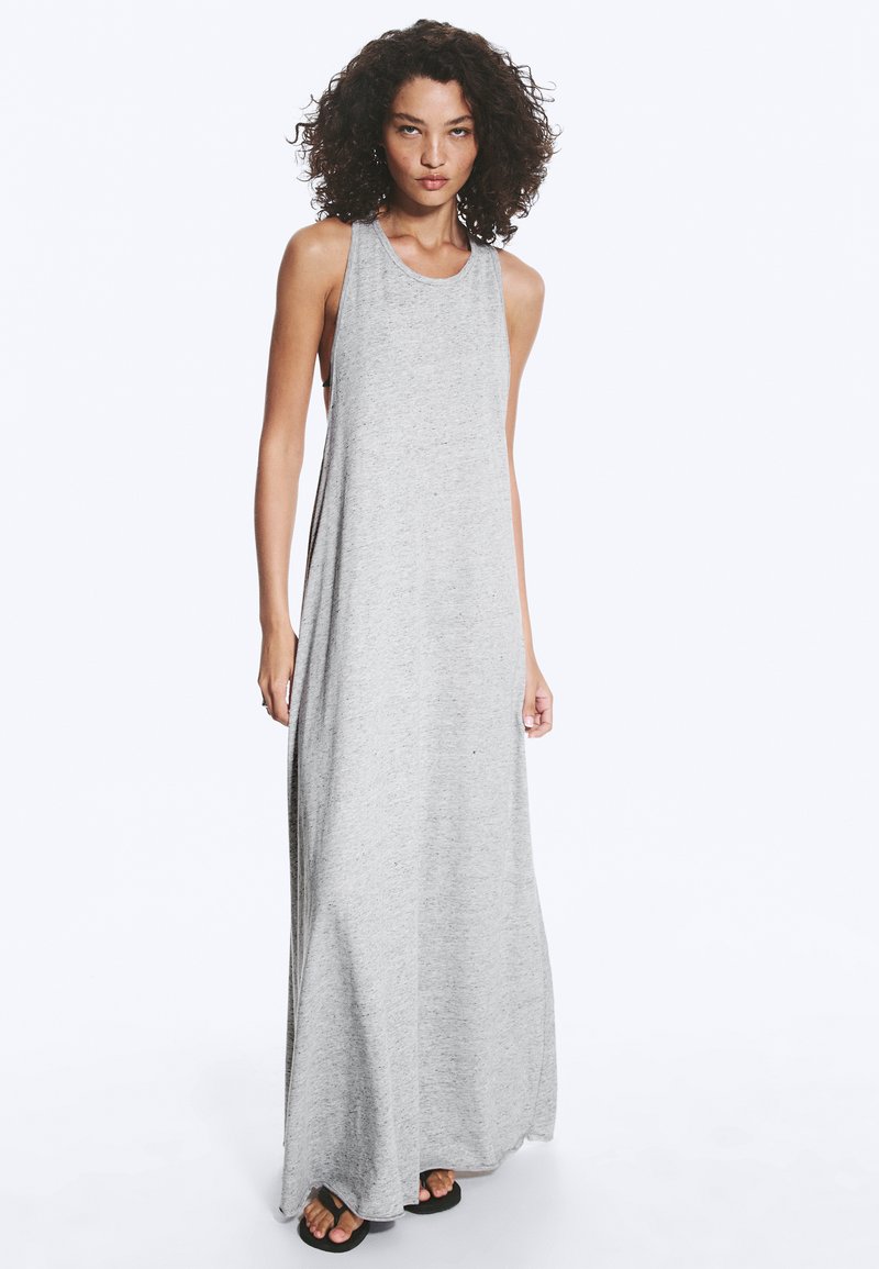 Femme aux cheveux bouclés portant une robe maxi gris clair sans manches et des sandales noires, debout devant un fond uni.