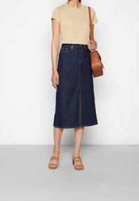 Denim-midi-kjol med främre slits, mörkblå färg och kontrasterande sömmar. Matchad med en beige t-shirt och en brun axelremsväska.