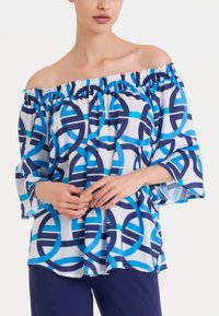 Blusa scollata realizzata in tessuto leggero con motivi geometrici blu e bianchi, dotata di un collo arricciato elasticizzato e maniche ampie.