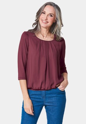 Blouse bordeaux avec un col rond, devant plissé et manches trois-quarts, associée à un jean bleu. Tissu doux et lisse.