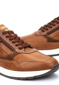Sneaker in pelle marrone con texture perforata, sovrapposizioni a contrasto e lacci marroni. Presenta una suola intermedia bianca e un dettaglio con zip laterale.