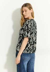 Sort bluse med hvide abstrakte mønstre, korte ærmer og en løs pasform, parret med blå jeans. Glat tekstur og afslappet design.