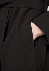 Vêtement noir avec une texture douce, présentant un détail de poche cousue. Le tissu a un léger éclat et une coupe ajustée.