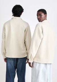 Crèmekleurige fleece pullover met een raglanmouw ontwerp en knoopsluitingen bij de mouwen, gecombineerd met een blauwe spijkerbroek en een lichtgekleurde rok.