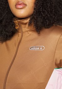 Close-up of a person wearing a brown Adidas zip-up jacket with logo on chest and curly dark hair visible.