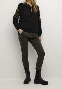 Haut à manches longues noir avec broderie en feuilles d'or, associé à un pantalon skinny vert olive avec des poches cloutées, et des bottines noires.