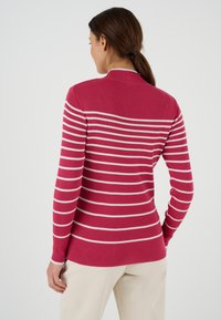 Donna con capelli castani raccolti in una coda di cavallo, che indossa un maglione a righe rosse e bianche a costine, in piedi con la schiena rivolta in avanti e pantaloni beige.