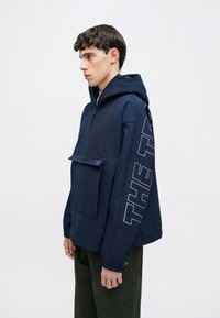 Sötétkék kapucnis dzseki elülső cipzárral, nagy első zsebbel és feltűnő szöveges grafikával az ujjon. Simafonás, könnyű anyagból készült.