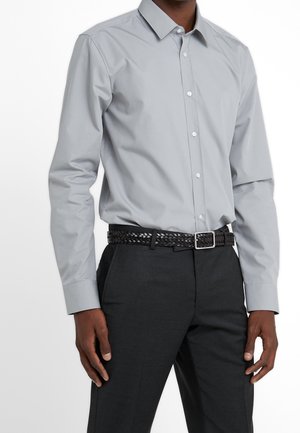 Man draagt een lichtgrijs overhemd met lange mouwen, ingestopt in donkere grijze broek met een zwarte gevlochten riem.