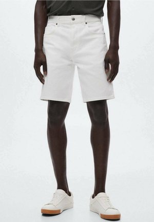 Weiße Jeans-Shorts mittlerer Länge, getragen von einer stehenden Person mit dunkler Haut, kombiniert mit weißen Sneakers und einem schwarzen, in das Hemd gesteckten Shirt.