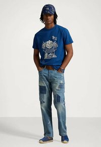 Άνδρας που φοράει μπλε γραφικό T-shirt, μ patched μπλε τζιν, μπλε καπέλο με σχέδιο και μπλε παπούτσια, στέκεται με τα χέρια στις τσέπες.