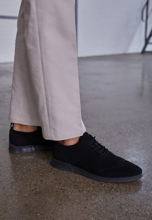 Personne portant un pantalon beige et des chaussures à lacets en maille noire debout sur un sol en béton poli tacheté.