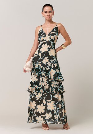 Frau trägt ein schwarzes Maxikleid mit beigen Blumenmuster, gestuften Rüschen, goldarmband, passenden Ohrringen, beiger Clutch und Sandalen.