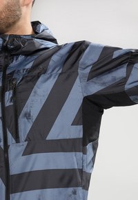 Veste extérieure pour hommes présentant un motif géométrique bleu et noir, fabriquée en matériau imperméable avec une fermeture éclair à l'avant et des accents en mesh.