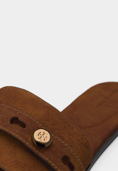 Sandale mule en daim marron avec bride réglable ornée d'un bouton décoratif rond en métal doré et de découpes en forme de serrure.