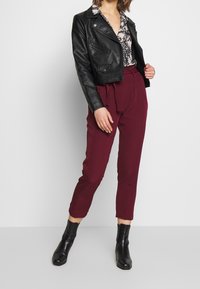 Schwarze Lederbikerjacke, gemusterte Bluse, bordeauxfarbene tapered Hose mit Krawattenbund und schwarze Knöchelstiefel. Glatte Texturen und taillierte Passform.