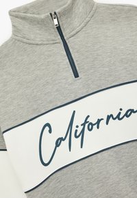 Grijze fleece pullover met een hoge kraag, voorzien van een halve rits. Een witte band over de borst toont "California" in marineblauw.