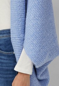 Jasnoniebieski szal z teksturą wykonany z miękkiego materiału, opadający na białą koszulę z długim rękawem i mankietem, w połączeniu z ciemnymi dżinsami.