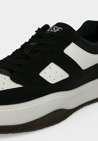 Baskets noir et blanc avec une semelle blanche épaisse, des lacets noirs et des accents blancs perforés sur le côté et la zone des orteils.
