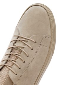 Sneaker in suede beige con suola in gomma, punta rotonda, lacci piatti e pannello elastico laterale. Presenta dettagli cuciti e una texture liscia.