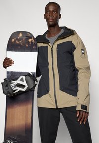 Σνόουμπορντερ με μπεζ και μαύρο μπουφάν, κρατώντας ένα σνόουμπορντ με λευκή και σκούρα κορυφή, με μαύρη δέστρα προσαρτημένη.