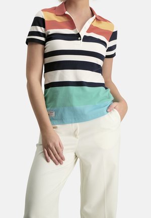 Frau trägt ein kurzärmeliges Poloshirt mit horizontalen Streifen in Rost, Gelb, Schwarz, Weiß, Grün und Blau, kombiniert mit einer weißen Hose.