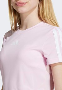 Light pink athletic shirt with short sleeves, featuring white striped accents on shoulders and a reflective logo on the front. Smooth texture.