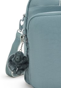 Shoulder bag in soft blue fabric, featuring a front zip pocket, adjustable strap, and a gray plush gorilla keychain attached.