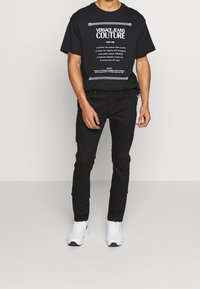 Svart bomullströja med vitt grafiskt tryck; kombinerad med svarta slim-fit byxor. Modellen bär vita sneakers med strukturerade sulor.